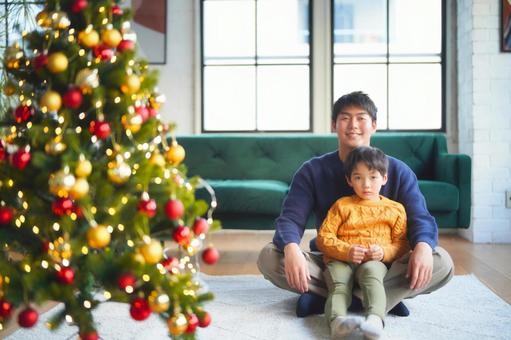 クリスマスツリーの横にあぐらで座る男性の上に足を伸ばして座る男の子 クリスマスツリーの横にあぐらで座る男性の上に足を伸ばして座る男の子 親子,男の子,小学生の写真素材