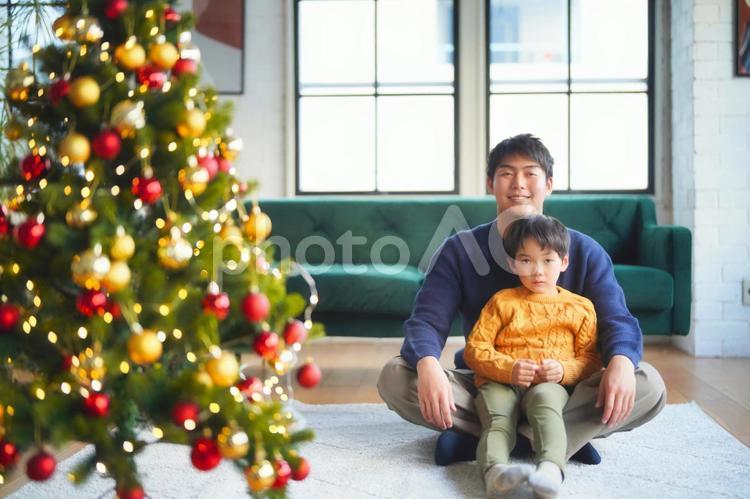 クリスマスツリーの横にあぐらで座る男性の上に足を伸ばして座る男の子 親子,男の子,小学生の写真素材