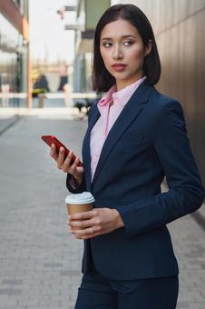 街なかでコーヒーとスマートフォンを持って立っているスーツの女性 ビジネスウーマン,ビジネス,女性の写真素材