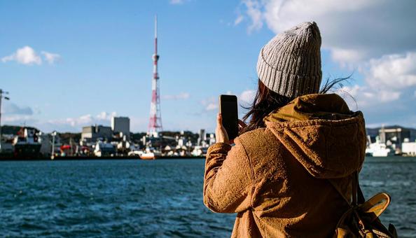 海の向こうを携帯で撮影する観光客の女性の写真