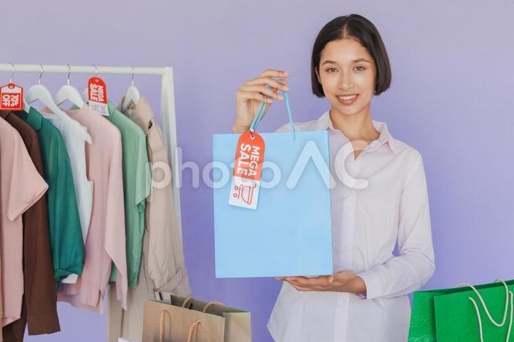 セールのタグの付いたハンガーに掛かった洋服と紙袋を持つカメラ目線のアジア人女性 セール,ショップ,女性の写真素材