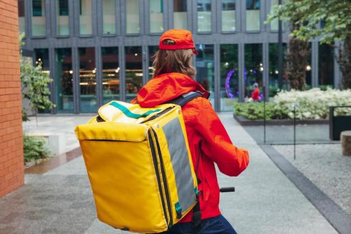 街なかにいる電動キックボードに乗るフードデリバリーの男性の後ろ姿の写真