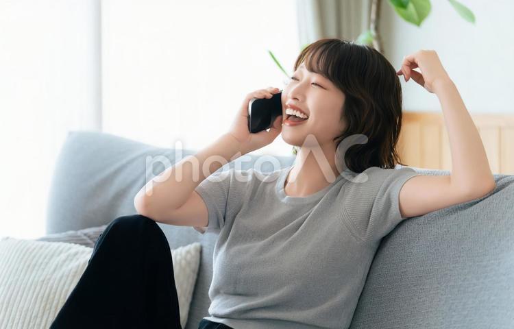 ソファに座って笑いながらスマホで話す女性 女性,スマホ,笑顔の写真素材