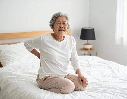 ベットの上で腰を抑えて苦しむ白髪の女性 ベットの上で腰を抑えて苦しむ白髪の女性の写真