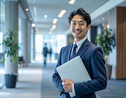 オフィスでPCを持って微笑む男性社員 オフィスでPCを持って微笑む男性社員の写真