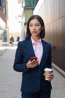 街なかでスマートフォンとコーヒーを持って立つスーツのアジア人女性 ビジネスウーマン,ビジネス,女性の写真素材