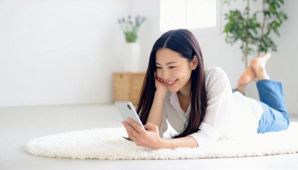 リビングで横になりスマホを操作する女性 リビングで横になりスマホを操作する女性の写真
