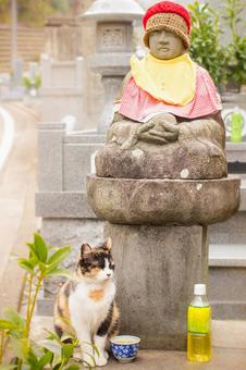 お地蔵さんの傍らに座る三毛猫の写真