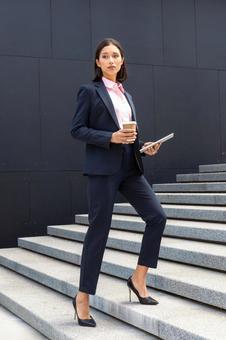 コーヒーとタブレットを持ってオフィスビルの外の階段を登るパンツスーツの女性 ビジネスウーマン,ビジネス,女性の写真素材