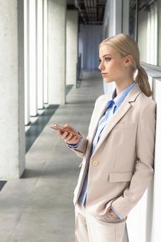 オフィスの通路の壁にもたれてスマートフォンを持って立つビジネスウーマン ビジネスウーマン,ビジネス,女性の写真素材
