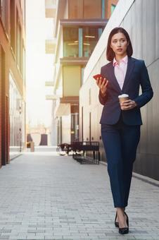 街なかでコーヒーとスマートフォンを持って歩いているビジネスウーマン ビジネスウーマン,ビジネス,女性の写真素材