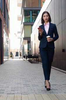 街なかでコーヒーとスマートフォンを持って歩くビジネスウーマン ビジネスウーマン,ビジネス,女性の写真素材