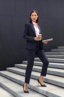 コーヒーとタブレットを持ってオフィスビルの外の階段を登るパンツスーツのビジネスウーマン ビジネスウーマン,ビジネス,女性の写真素材