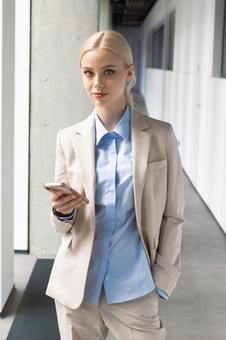 オフィスの通路でスマートフォンを持って立つカメラ目線のビジネスウーマン ビジネスウーマン,ビジネス,女性の写真素材