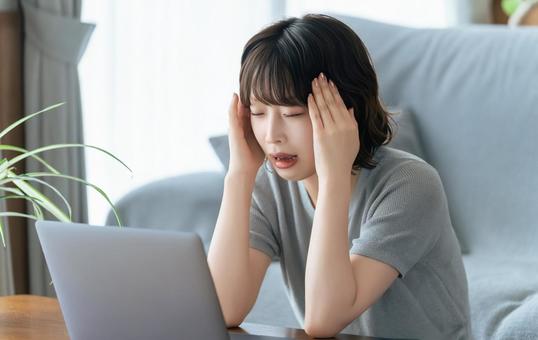 パソコンを使いながら頭を押さえて悩む女性 パソコンを使いながら頭を押さえて悩む女性の写真