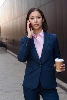 街なかでコーヒー片手に電話しているパンツスーツの女性 ビジネスウーマン,ビジネス,女性の写真素材