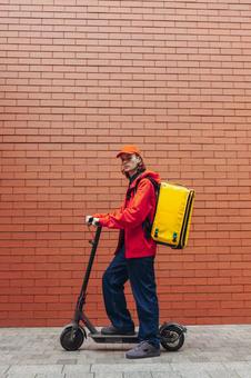 レンガ壁の前で電動キックボードに乗るカメラ目線の配達員の男性 配達,フードデリバリー,デリバリーの写真素材