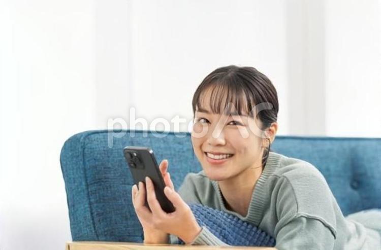ソファで寝転んでスマートフォンを見る女性 女性,スマートフォン,ソファの写真素材