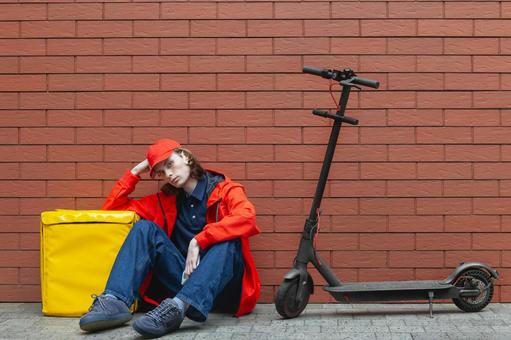 電動キックボードの横に座り込んでいるフードデリバリーの男性 配達,フードデリバリー,デリバリーの写真素材