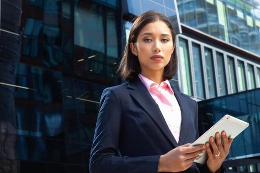 オフィスビルの外でタブレットを持ってカメラ目線で立っているスーツのアジア人女性 ビジネスウーマン,ビジネス,女性の写真素材