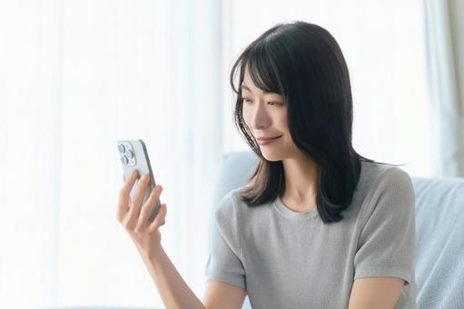 ソファに座ってスマホを使う笑顔の女性 ソファに座ってスマホを使う笑顔の女性の写真