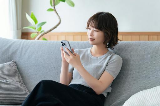 ソファに座ってスマホを使う笑顔の女性 ソファに座ってスマホを使う笑顔の女性の写真