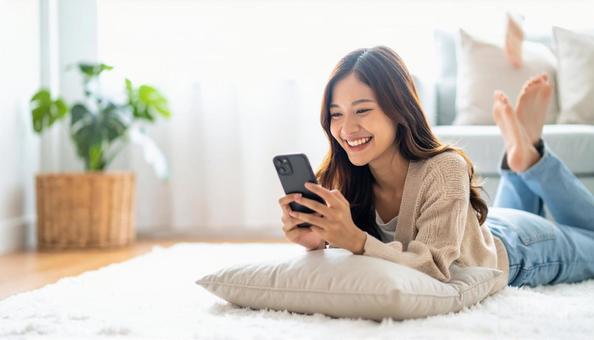 リビングで横になりスマホを操作する女性 リビングで横になりスマホを操作する女性の写真