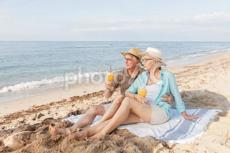 海辺に敷物を引いてジュースを持って遠くを見るシニアカップル 男性,女性,男女の写真素材