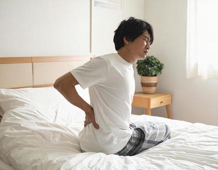 ベットの上で腰を抑えて苦しむ男性 ベットの上で腰を抑えて苦しむ男性の写真