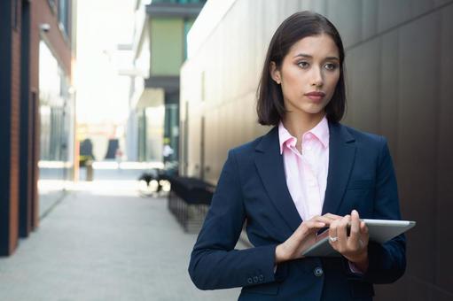 街なかでタブレットを持って立つスーツの女性 ビジネスウーマン,ビジネス,女性の写真素材