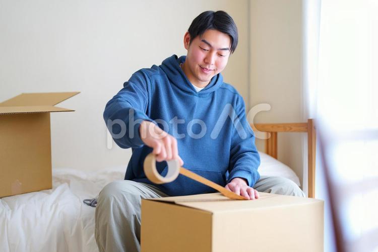 ベッドに座って段ボール箱の蓋をテープで留める男性 引っ越し,一人暮らし,男性の写真素材