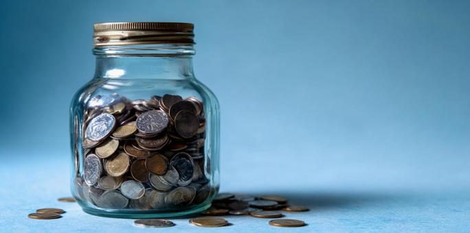青い背景に輝く、コインが詰まった貯金瓶の写真