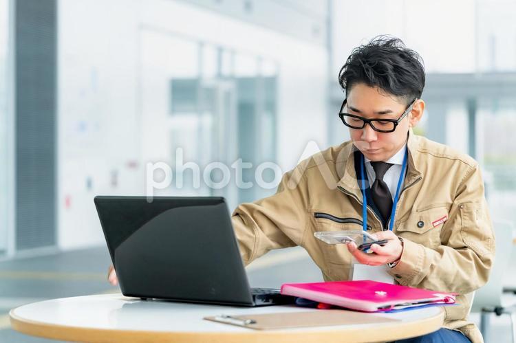 携帯を見ながらパソコンを触る作業服の男性 作業服,ビジネス,ビジネスマンの写真素材