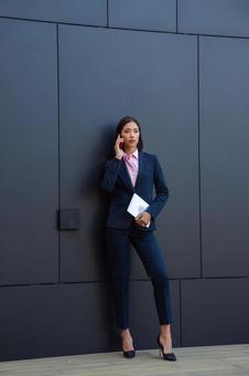 オフィスビルの黒い壁の前で電話しているパンツスーツのアジア人ビジネスウーマン ビジネスウーマン,ビジネス,女性の写真素材
