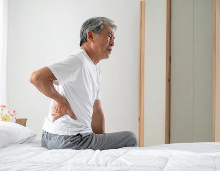 ベットの上で腰を抑えて苦しむシニア男性 ベットの上で腰を抑えて苦しむシニア男性の写真