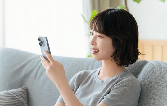 ソファに座ってスマホを使う笑顔の女性 ソファに座ってスマホを使う笑顔の女性の写真