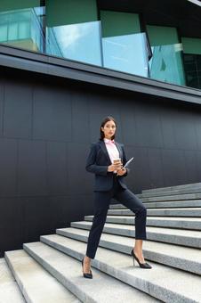 コーヒーとタブレットを持ってオフィスビルの外の階段を登っているパンツスーツの女性 ビジネスウーマン,ビジネス,女性の写真素材