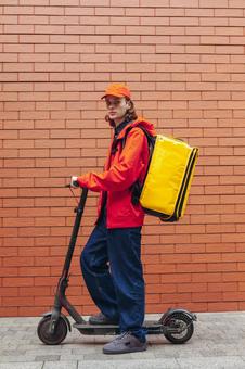レンガ壁の前で電動キックボードに乗るカメラ目線のフードデリバリーの男性 配達,フードデリバリー,デリバリーの写真素材