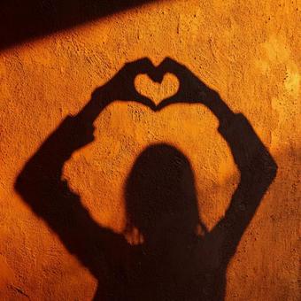 オレンジの壁に映る手で作ったハートの影の写真