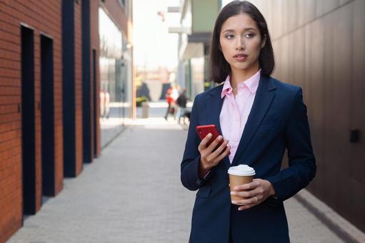 街なかでスマートフォンとコーヒーを持って立つアジア人ビジネスウーマン ビジネスウーマン,ビジネス,女性の写真素材