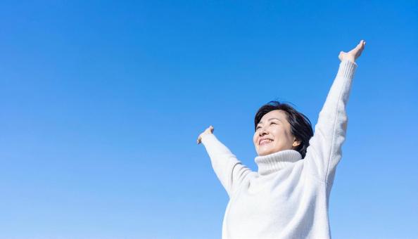 青空の下で伸びをする秋服の日本人女性の写真