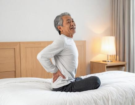 ベットの上で腰を抑えて苦しむシニア男性の写真